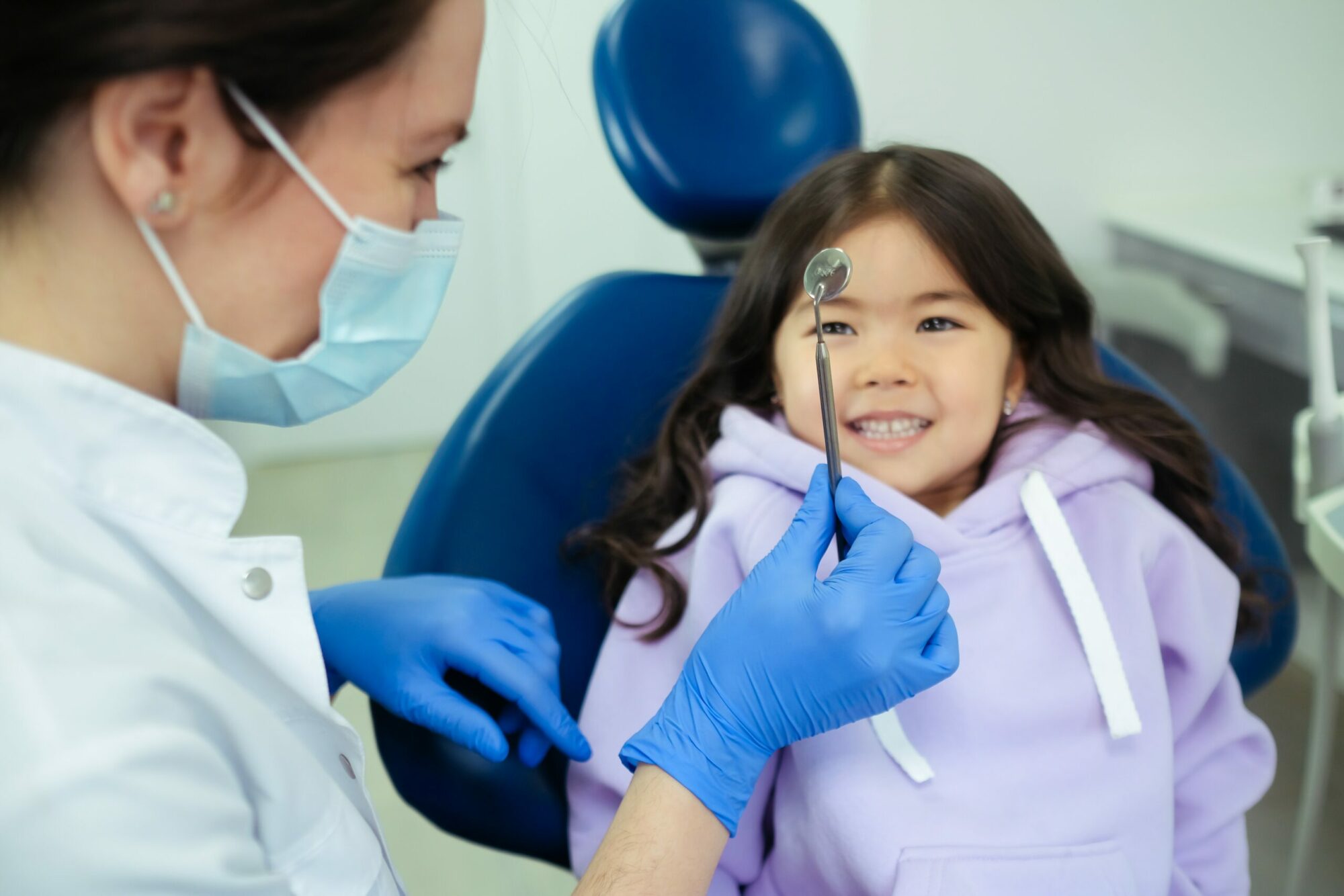 a child with long hair is sitting in a dentist chair, smiling at the dentist who is holding up a tool