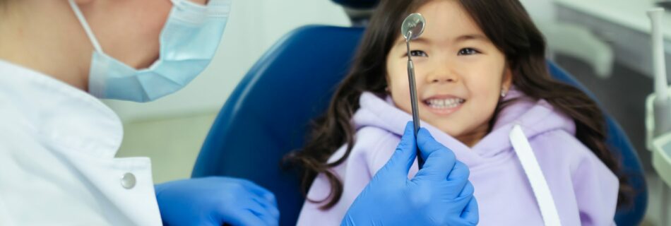 a child with long hair is sitting in a dentist chair, smiling at the dentist who is holding up a tool