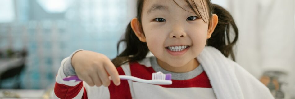 A young girl smiling as she holds out a purple tipped toothbrush.