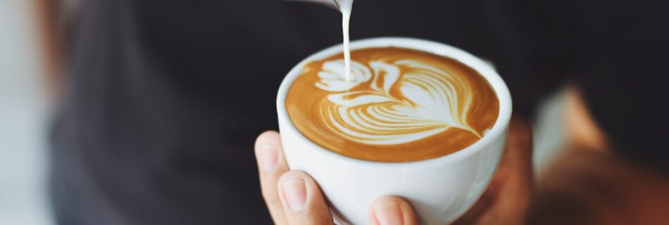 Person pouring a latte art of a flower in a white ceramic mug.