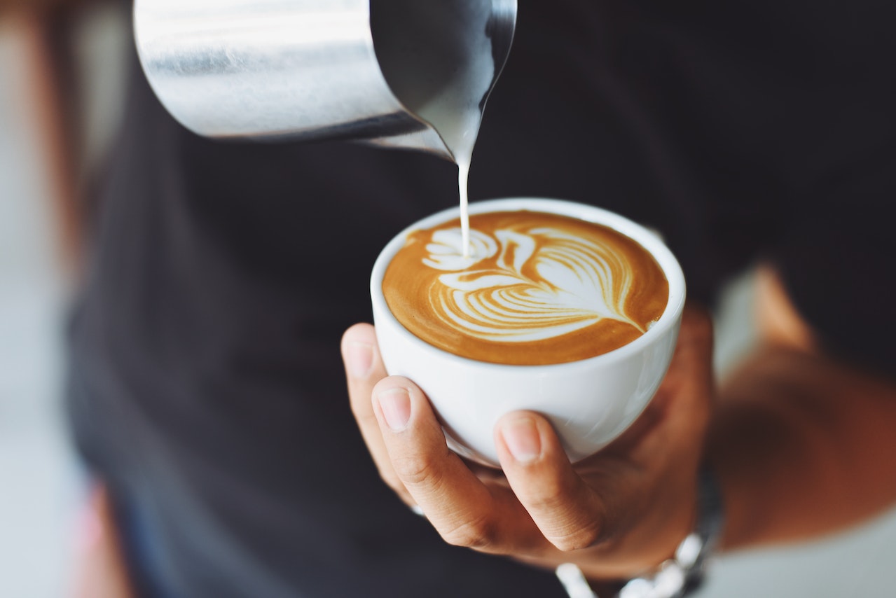Person pouring a latte art of a flower in a white ceramic mug.