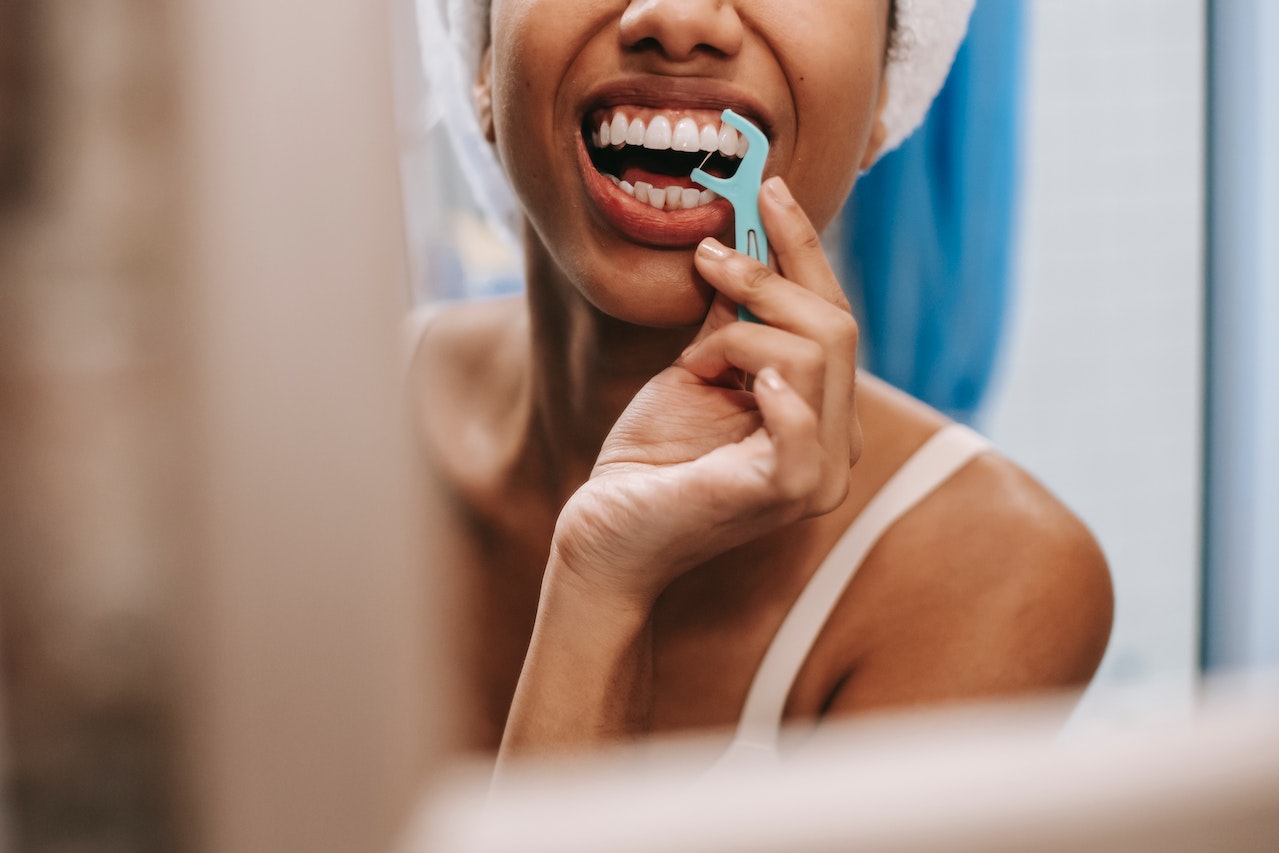 Reflection of a person with a towel on their head using a flossing stick to floss their teeth in a mirror.