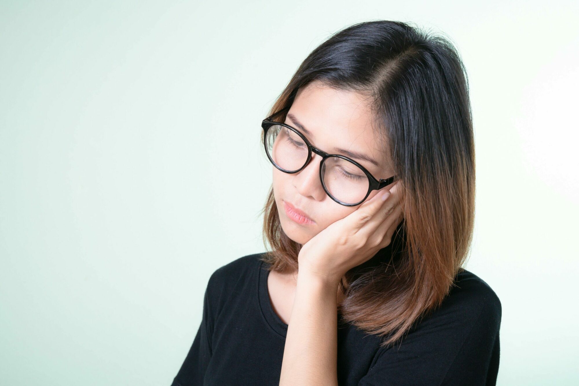 An Asian woman with straight dark brown hair and glasses standing with her head to the side, leaning her jaw into her hand