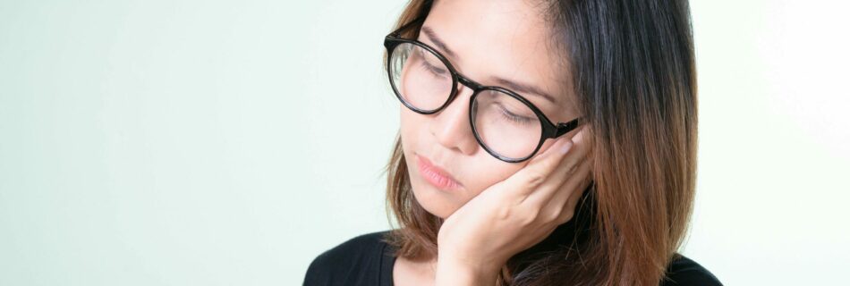An Asian woman with straight dark brown hair and glasses standing with her head to the side, leaning her jaw into her hand