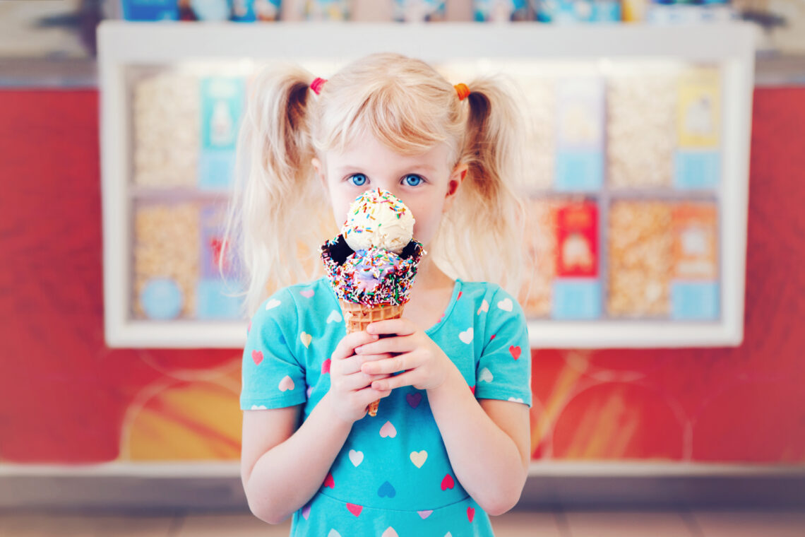 Cute adorable Caucasian blonde preschool girl child with blue eyes holding ice cream in large waffle cone with sprinkles. Kid with frozen food. Happy childhood lifestyle. Tasty summer dessert