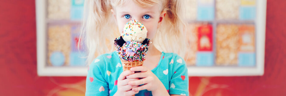 A young child is seen standing in front of an ice cream stand holding a giant ice cream cone in front of their face.