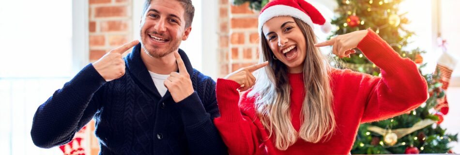 Young couple wearing santa claus hat sitting on chair and table around christmas tree at home smiling cheerful showing and pointing with fingers teeth and mouth. Dental health concept.